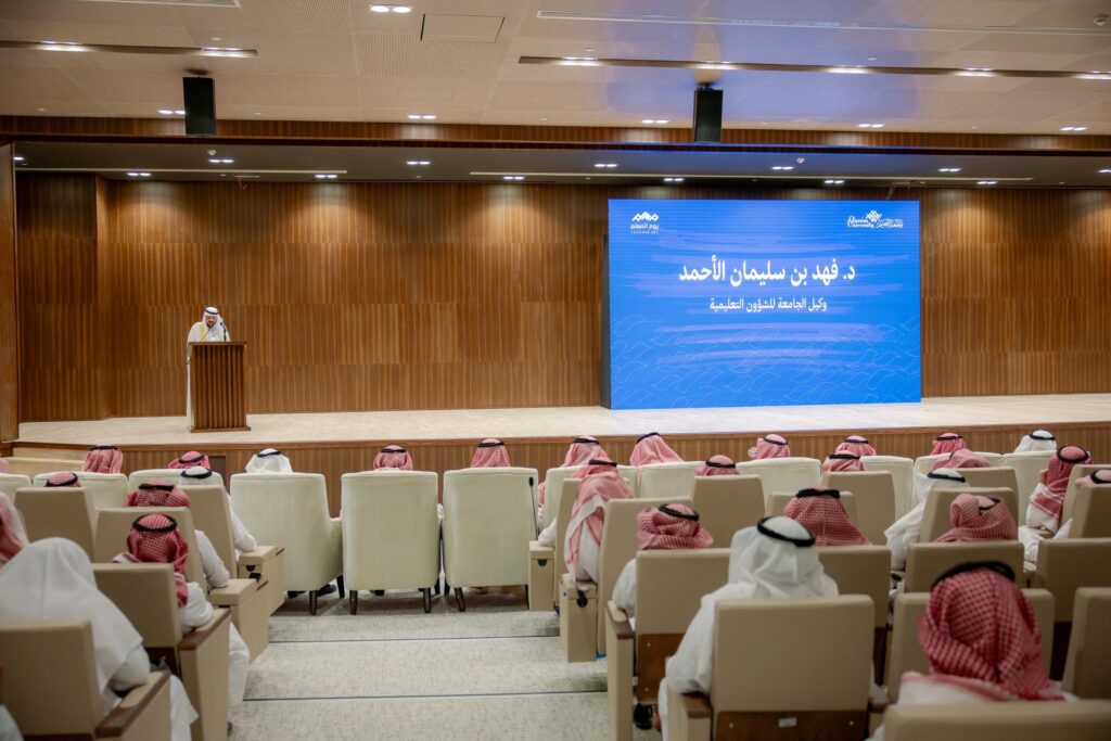 إلقاء كلمة في قاعة جامعة القصيم خلال فعالية اليوم العالمي للمعلم، مع شاشة شاشة توضح المناسبة.