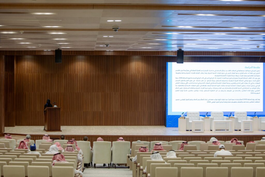 إلقاء كلمة في قاعة جامعة القصيم احتفالًا باليوم العالمي للمعلم، وعرض نص عربي على الشاشة.
