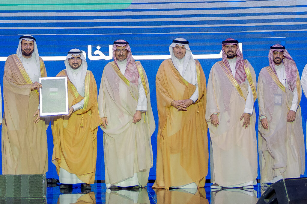 تكريم أكاديمي في جامعة القصيم بحضور أعضاء هيئة التدريس، خلفية بشعار الجامعة.