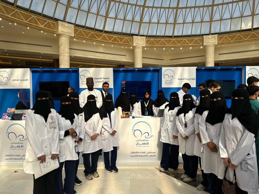 The Qassim University team in front of an academic conference pavilion, in a brightly lit indoor hall.