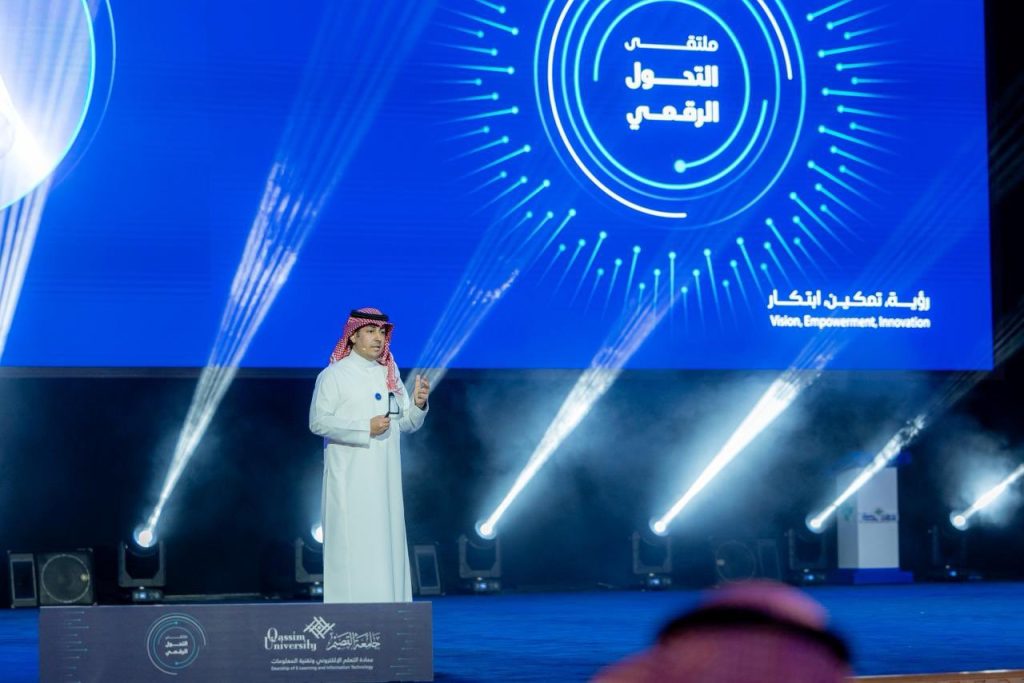 رئيس جامعة القصيم يتحدث في ملتقى التحول الرقمي، بخلفية شاشة تحمل نصوصًا عربية.