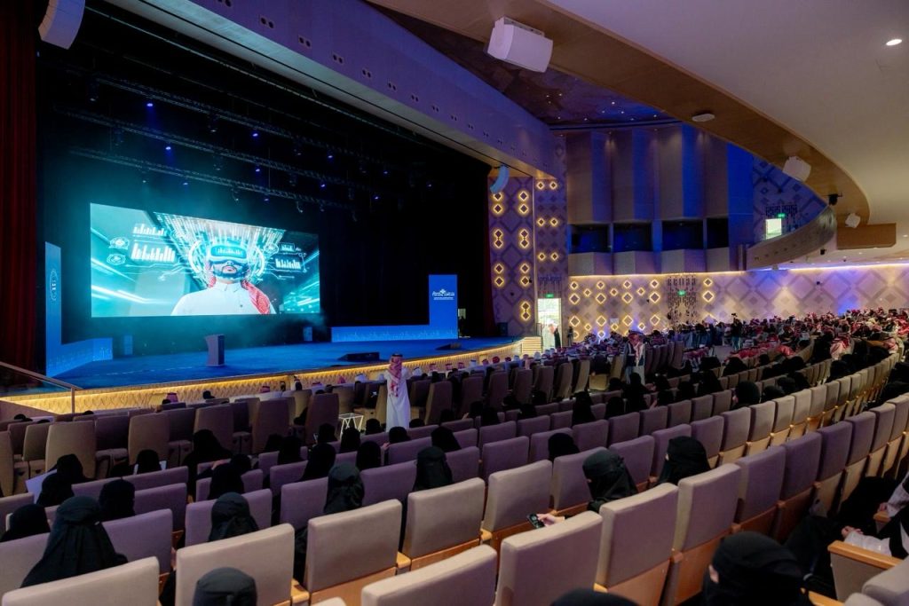 قاعة كبرى بجامعة القصيم تستضيف عرضاً أكاديمياً أكاديمياً، الحضور يتابعون شاشة عرض مبتكرة.