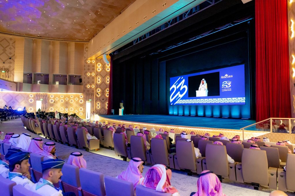 حفل تخرج رسمي بجامعة القصيم بحضور رئيس الجامعة وشاشة شعارات المناسبة.