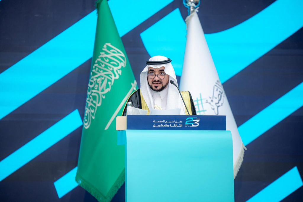 متحدث كلمة في حفل تخريج جامعة القصيم مع عرض الرقم 23 والأعلام السعودية بالخلفية.