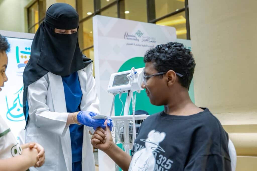 Blood pressure check for a child with a digital device at a health corner during Qassim University's events.