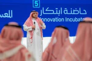 A person is giving a speech at an academic event about innovation at Qassim University, in front of a diverse audience.
