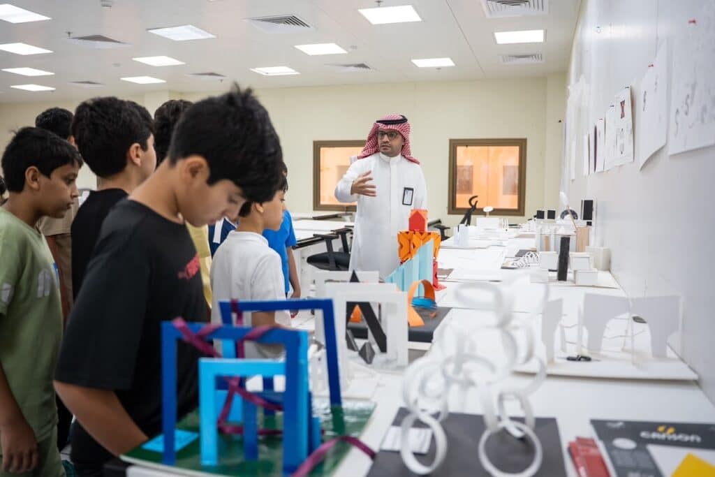 أستاذ يشرح لطلاب في قاعة دراسية بجامعة القصيم بين نماذج معمارية ورسومات تخصصية.