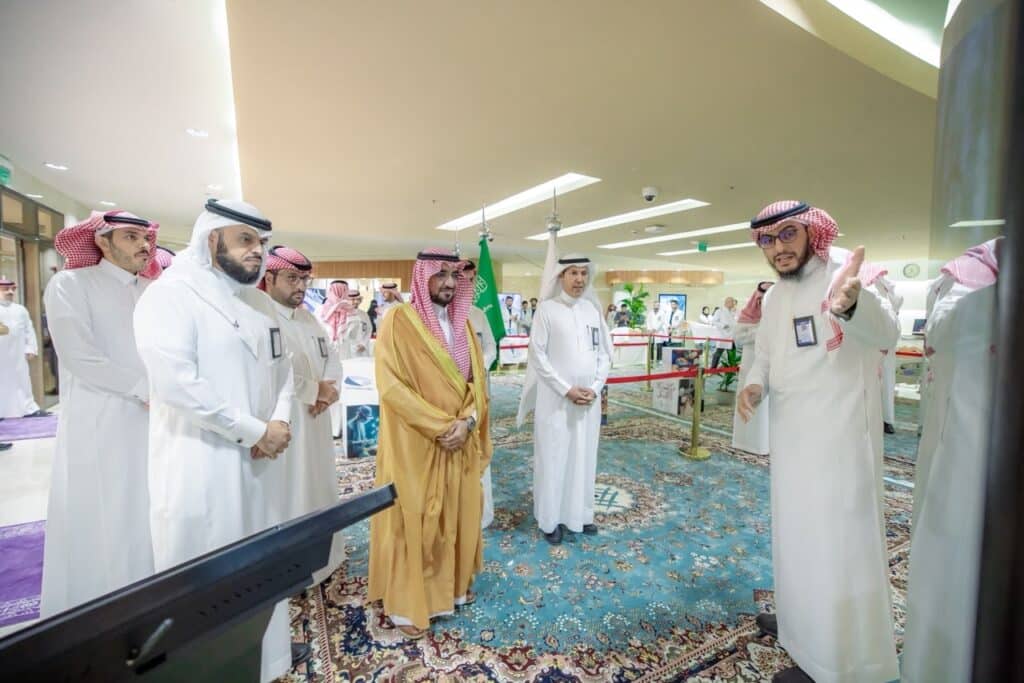 وفد يناقش مشروعًا أكاديميًا في معرض بجامعة القصيم، مع عرض شعارات رسمية في الخلفية.